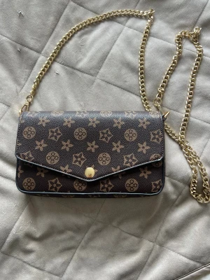 Brun handväska med kedjerem - Snygg brun handväska med guldigt kedjeband och tryckknapp. Väskan har ett klassiskt mönster i beige och brunt samt matchande plånbok. Tillverkad i konstläder med stilren design och praktisk storlek, perfekt för att bära det viktigaste.