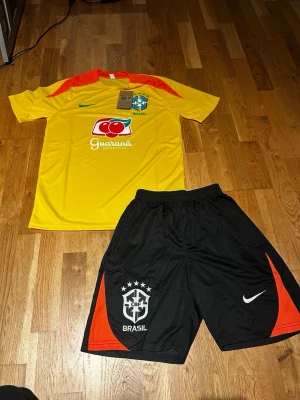 Brazil summer set Yellow/black - Säljer ett Brasilien fotbollskit från Nike med gul t-shirt och svarta shorts. Tröjan har röda och gröna detaljer, tryckt logga och sponsortryck. Shortsen har Brasilien-emblem och röda detaljer på sidorna. Perfekt för fotbollsträning eller match!
