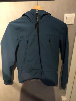 Blå vindjacka med huva från C.P. Company - Snygg blå vindjacka från C.P. Company med dragkedja snett framtill, två fickor med dragkedja och ikoniska glasögon i huvan. Jackan har en sportig look och är tillverkad i ett lätt, vindtätt material. Perfekt för dig som gillar streetwear och funktionella detaljer.PRISET KAN DISKUTERAS!