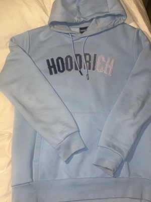 Ljusblå hoodie från Hoodrich - Snygg ljusblå hoodie från Hoodrich med broderad logga i mörkblått och vitt på bröstet. Klassisk modell med huva och känguruficka framtill. Perfekt för en avslappnad streetwear-look.