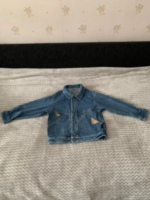Blå jeansjacka med triangelfickor - Snygg blå jeansjacka med klassisk krage och unika triangelformade fickor framtill. Jackan har knäppning med metallknappar och långa ärmar med knapp vid ärmslut. Perfekt för dig som vill ha en cool och avslappnad stil.