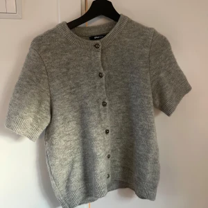 Grå kortärmad kofta från Gina Tricot  - Säljer en ljusgrå, stickad kofta från Gina Tricot med korta ärmar och rund halsringning. Koftan har knappar framtill och ribbade kanter vid ärmslut och nederkant.