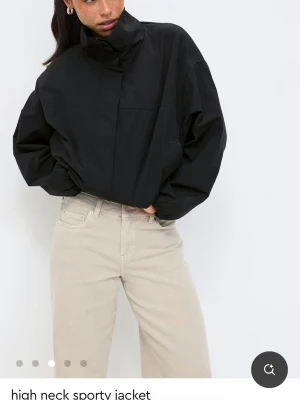 Svart vindjacka med hög krage - Stilren svart vindjacka med hög krage och oversized passform. Jackan har dold dragkedja framtill och är tillverkad i ett lätt, syntetiskt material som passar perfekt för blåsiga dagar. Enkel och sportig design som funkar till det mesta.