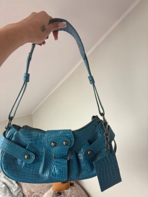 Blå axelväska i krokopräglat mönster - Säljer en blå handväska i fejkat krokodilskinn med justerbart axelband och silverfärgade detaljer. Väskan har en unik form, dragkedja upptill och dekorativa spännen framtill. Perfekt för dig som gillar statement-accessoarer och trendig design.