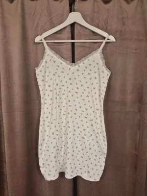 Vit blommig klänning med spets - Söt vit klänning med små blommor i rosa och grönt. Klänningen har smala axelband, v-ringad spetskant och ärmlös design. Ribbad struktur ger en trendig känsla och den är perfekt för sommaren.