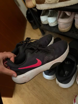 Svarta Nike sneakers med rosa swoosh - Snygga svarta Nike sneakers med en tydlig rosa swoosh på sidan. Skorna har en vit och svart sula med bra grepp och ovandel i meshmaterial för bra ventilation. Klassisk låg modell med snörning, perfekt för en sportig och avslappnad stil.