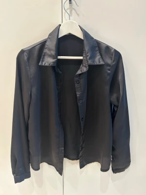 Svart långärmad skjorta i satin - Stilren svart skjorta i glansigt satinmaterial med klassisk krage och långa ärmar. Skjortan har knappar framtill och en rak passform som ger en clean look. 