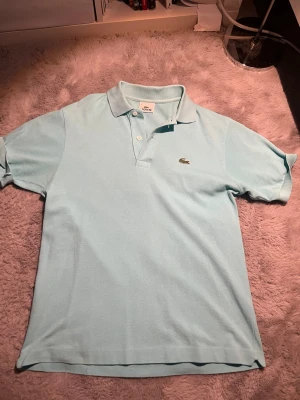 Ljusblå pikétröja från Lacoste - Snygg ljusblå pikétröja från Lacoste med klassisk krage och två knappar framtill. Tröjan har det ikoniska krokodilmärket broderat på bröstet och är tillverkad i mjuk bomull. Perfekt för en clean och fräsch look.