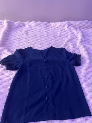 Mörkblå blus med spetsdetaljer - Söt mörkblå blus med korta ärmar och spetsdetaljer vid ärmsluten. Blusen har vågig knappslå framtill med matchande knappar och en mjuk, feminin siluett. Perfekt för dig som gillar stilrena men unika plagg.