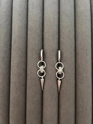 Silvriga örhängen med ringar och spets - Snygga silvriga örhängen med två sammankopplade ringar och en spetsig hängande detalj längst ner. Örhängena har en modern och edgy design som sticker ut. Perfekta för dig som gillar minimalistiska men ändå unika accessoarer.