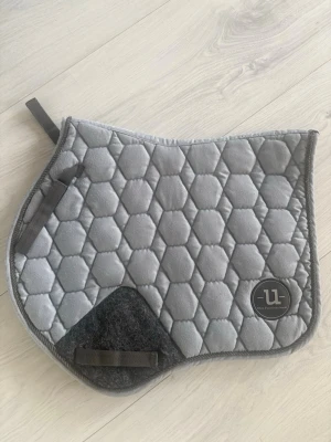 Grått schabrak från Uhip - Säljer ett grått schabrak från Ulla med sexkantigt quiltat mönster och förstärkt svart filtparti. Schabraket har praktiska fästen och en rundad form som passar till sadel. Perfekt för dig som gillar stilrena hästtillbehör.