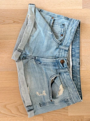 Ljusblå jeansshorts med slitningar - Säljer ett par ljusblå jeansshorts från Dr. Denim med uppvikta benslut och slitna detaljer framtill och baktill. Klassisk femficksmodell med knapp och dragkedja. Perfekta för sommaren och ger en avslappnad look.