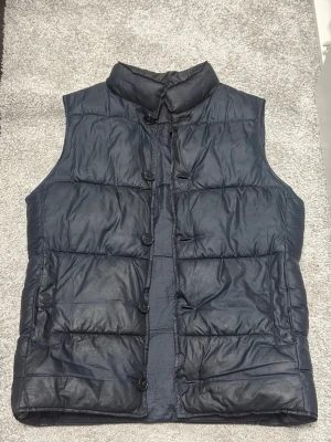 Mörkblå dunväst från John Henric - Snygg mörkblå dunväst med ståkrage och knäppning framtill med stora knappar. Västen har två framfickor och är quiltad för extra värme. Perfekt för lager-på-lager och enkel att matcha med olika outfits. Ha en skön varm vår!