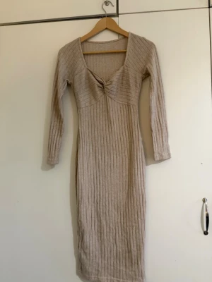 Beige ribbad långklänning - Beige ribbad långklänning med lång ärm och v-ringad halsringning med twistdetalj framtill. Klänningen har en kroppsnära passform och är tillverkad i ett stickat material som känns mjukt och följsamt.