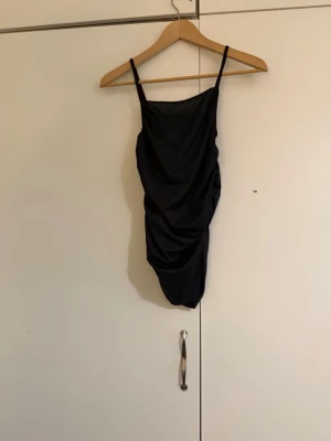 Svart linne med tunna axelband - Snygg svart body med tunna, justerbara axelband och draperad effekt på sidan. Tillverkad i ett stretchigt material som ger en smickrande passform. Perfekt att styla med jeans eller kjol för en trendig look.