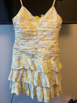 Gul volangklänning med mönster - Supersöt kort klänning i gult med vitt paisleymönster och volanger. Klänningen har v-ringning, smala axelband och flera lager volanger nedtill för en extra flowy look. Perfekt för sommarens alla tillfällen! Köpt för ett år sen och har använts ett par gånger så klänningen är i väldigt bra skick❣️