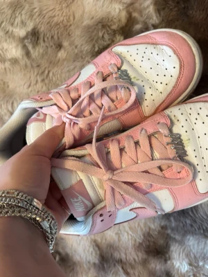 Nike Dunk Low rosa och vita sneakers - Nike Dunk Low sneakers i vitt och ljusrosa läder med rosa snörning och gummisula. Klassisk Nike-logga på sidan och tungan. Perforerad tå för extra ventilation. Skapad på Nike egna hemsida så färgen är personsanpassad och den silvriga detaljen vid skosnöret 