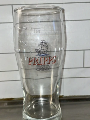 Pripps ölglas med skeppmotiv - Stort ölglas i genomskinligt glas med Pripps-logga och ett tryckt skeppsmotiv på framsidan. Glaset har en klassisk, avsmalnande form och är dekorerat med röda bokstäver och tre mynt under loggan. Perfekt för dryckesentusiasten.