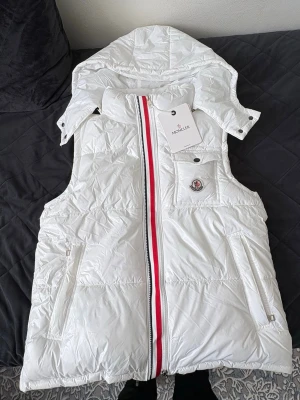 Vit dunväst från Moncler med huva - Snygg vit dunväst från Moncler med avtagbar huva och klassisk röd-vit-blå dragkedja. Västen har två sidofickor med dragkedja och en bröstficka med Moncler-logga. Materialet är glansigt och quiltat, perfekt för att hålla värmen. Cool detalj med Moncler-serie på insidan.