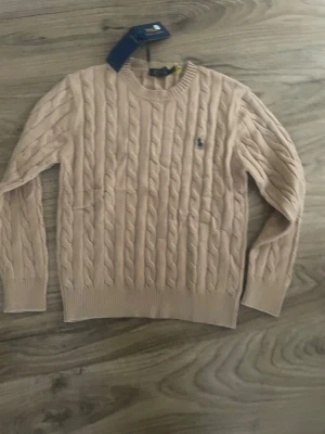 Beige kabelstickad tröja Polo Ralph Lauren - Snygg beige kabelstickad tröja från Polo Ralph Lauren med rund hals och diskret blå logga broderad på bröstet. Tröjan har ribbade muddar och är tillverkad i mjukt material som passar perfekt för lager-på-lager. Klassisk och tidlös design.