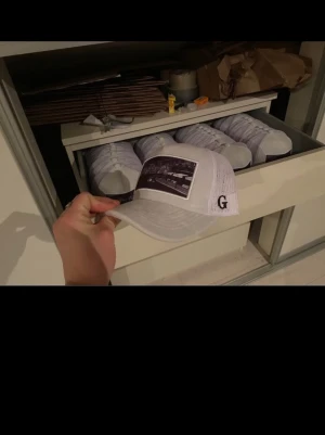 Vit keps med svart tryck och G-logga - Säljer en vit keps med svart tryck framtill och en liten G-logga på sidan. Kepsen har klassisk böjd skärm och mesh-paneler bak för extra ventilation. Perfekt för dig som vill ha en stilren och modern accessoar.