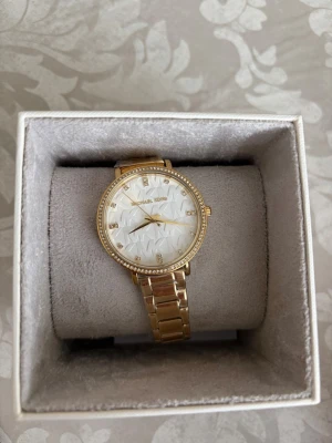 Guldig klocka från Michael Kors. Michael Kors Pyper - Snygg guldig klocka från Michael Kors med länkarmband och glittrande stenar runt urtavlan. Urtavlan är vit med ett geometriskt mönster och guldfärgade visare. Perfekt accessoar för att lyfta din stil. !!!Nypris:2200!! Pris kan diskuteras 