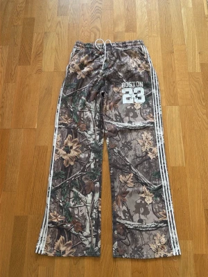 Camo mjukisbyxor med Boston-tryck - Säljer ett par camofärgade mjukisbyxor med vita ränder längs sidorna och trycket 'Boston 23' på ena benet. Byxorna har dragsko i midjan och avslappnad passform. Perfekta för dig som gillar streetwear och sportig stil.