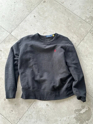 Svart sweatshirt från Polo Ralph Lauren - Klassisk svart sweatshirt från Polo Ralph Lauren med rund halsringning och diskret röd logga broderad på bröstet. Tröjan har långa ärmar och mjukt material på insidan för extra komfort. Perfekt till jeans eller joggers.