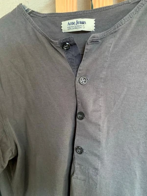 Acne Studios tröja - Rak, tunn, långärmad tröja i storlek S från Acne. 100% bomull, knäppning vid kragen, långärmad med muddar. Färgen är mörkgrå/mörkgrön. Litet hål på baksidan av ena ärmen, knappt märkbart (se bild), samt skada på mudd. Well-worn, vilket går ihop med tröjans stil!