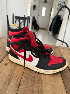 Air Jordan 1  - Säljer ett par Air Jordan 1 Retro High sneakers i klassisk röd, svart och vit färgkombination. Skorna har svart läder runt tån och hälen, röd tåbox och plös samt vit sidopanel med röd swoosh. Ikonisk vinge-logga på sidan och svart snörning.     Finns tecken på användning därför ett bra pris