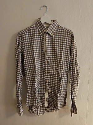 Brun och vit rutig skjorta - Klassisk brun och vit rutig skjorta med knappar framtill och lång ärm. Skjortan har en tidlös design och passar perfekt till jeans eller chinos. Tillverkad i mjuk bomull för en bekväm känsla.