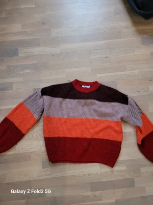 Stickad tröja från NA-KD - Färgglad stickad tröja från NA-KD i storlek M. Tröjan har breda ränder i vinrött, orange, rosa och rött. Den har en ribbad rund halsringning och lådformad passform med ballongärmar. Perfekt för dig som gillar retrovibbar.