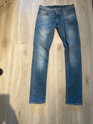 Nudie jeans - Säljer ett par blå slim fit jeans med snygga slitningar och klassisk femficksdesign. Jeansen har normal midja och smal passform hela vägen ner. Perfekta till en avslappnad stil och passar till det mesta. Materialet är denim i bomull.