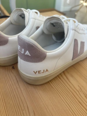Vita Veja sneakers  - Snygga vita sneakers från Veja med rosa mockadetaljer på häl och sidor. Klassisk låg modell med Veja-logga i beige på sidan och bak.Väldigt bra skick men de är lite smutsiga på vissa ställen men det tänker man inte på💗