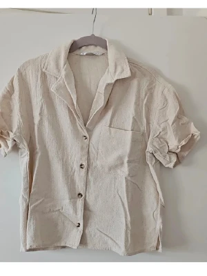 Beige kortärmad linneskjorta - Säljer en beige skjorta i linne med korta ärmar och klassisk krage. Skjortan har knappar framtill och en bröstficka. Perfekt för varma dagar och enkel att styla till olika outfits.