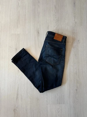 Mörkblå slim fit jeans från Jack & Jones - Snygga mörkblå slim fit jeans från Jack & Jones med klassisk femficksdesign och läderpatch bak i midjan. Jeansen har lätt slitna detaljer och raka ben. Perfekta för en avslappnad och trendig look.