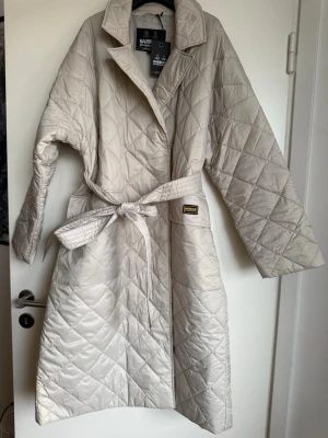 Ljusbeige quiltad kappa Barbour - Snygg ljusbeige quiltad kappa från Barbour International med bälte i midjan och klassisk krage. Kappan har ett rutmönster, långa ärmar och en stilren look. Perfekt för dig som vill ha en trendig och bekväm ytterjacka i lätt material.