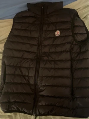 Svart dunväst från Moncler - Snygg svart dunväst från Moncler med klassisk quiltad design och hög krage. Västen har dragkedja framtill och Monclers ikoniska logotyp på bröstet. Perfekt för lager-på-lager och håller dig varm med stil.