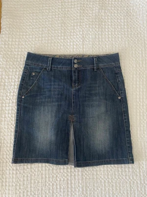 Blå y2k jeanskjol - Snygg blå jeanskjol i klassisk modell med två knappar i midjan, fickor fram och slits framtill. Pris kan diskuteras