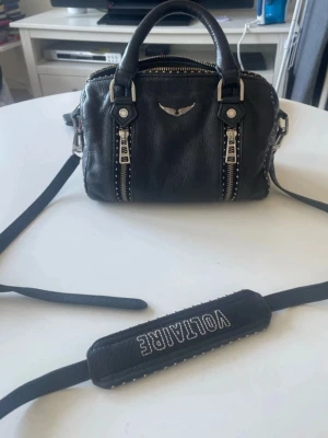 Svart Zadig & Voltaire handväska - Svart handväska i läder från Zadig & Voltaire med silverfärgade detaljer, dubbla dragkedjor framtill och nitar längs kanterna. Har tyvärr inget äktihetsbevis men den är äkta. 🌸Modellen är mellan
