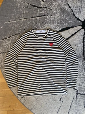 Randig långärmad topp Comme des Garçons - Säljer en svartvit randig långärmad topp från CDG Comme des Garçons PLAY med det ikoniska röda hjärtat på bröstet. Klassisk rund halsringning och mjukt bomullsmaterial. Perfekt för dig som gillar streetstyle och vill ha något stilrent men ändå lekfullt. || Strolek M ||