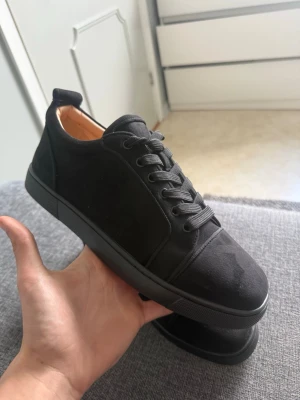 Svarta Louboutin sneakers i mocka - Snygga svarta sneakers från Christian Louboutin i mocka med klassisk röd sula. Skorna har lågt skaft, svarta skosnören och diskret branding på hälen. Perfekta för dig som gillar stilrena och exklusiva sneakers med en lyxig touch. Sprillans nya!!! Skickar inom 1-5 timmar låda också  inga byten inte prut bart
