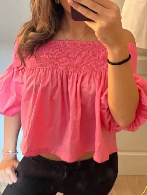 Rosa offshoulder blus med puffärmar - Trendig rosa offshoulder blus med bred smock upptill och voluminösa puffärmar. Luftig och croppad modell som ger en avslappnad men snygg look. Perfekt för dig som vill sticka ut med färg och stil.