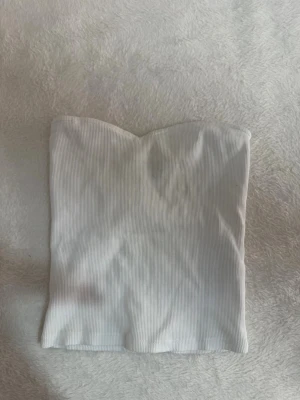 Vit ribbad bandeau tubtopp - Snygg vit bandeau tubtopp i ribbat material. Toppen är axelbandslös och har en lätt hjärtformad kant upptill. Perfekt att styla med jeans eller kjol för en trendig look.