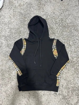 Svart Burberry hoodie  - Nästan helt ny och använd 1-2 gånger. Snygg svart hoodie från Burberry med klassiska beige-rutiga paneler längs ärmar och axlar. Stor huva med dragsko och Burberry-logga på snörena. Mjuk bomullskänsla och känguruficka framtill. Perfekt för en trendig streetwear-look.