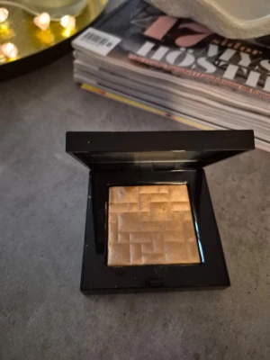 Bobbi Brown guld highlighter - Säljer en highlighter från Bobbi Brown i en lyxig svart dosa. Pudret har en skimrande guldton och en snygg geometrisk mönstring i ytan. Perfekt för att ge glow till ansiktet och framhäva kindbenen. Kompakt och enkel att ta med i väskan. Testad, swachad, fint skick., färg moon glow. 