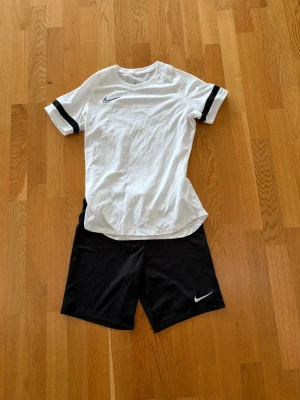 Nike Dri-Fit fotbollskit svart/vit - Nike Dri-Fit fotbollskit med vit t-shirt och svarta shorts. T-shirten har svarta detaljer på ärmarna och båda plaggen har Nike-logga. Tillverkad i funktionsmaterial som andas, perfekt för träning eller match.