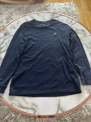 Marinblå långärmad topp Lyle & Scott - Snygg marinblå långärmad topp från Lyle & Scott Junior med klassisk rund hals och gul logga broderad på bröstet. Tillverkad i mjuk bomull som känns skön mot huden. Perfekt för en avslappnad och stilren look.