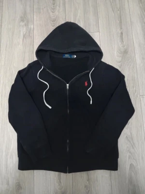 Svart zip hoodie från Polo Ralph Lauren - Svart hoodie med dragkedja från Polo Ralph Lauren. Storlek L men sitter som M. Klassisk design med vit dragsko i huvan och röd broderad logga på bröstet. Tillverkad i mjukt bomullsmaterial, perfekt för en avslappnad stil. Nypris 2999 mitt pris 599. De är dock två hål.