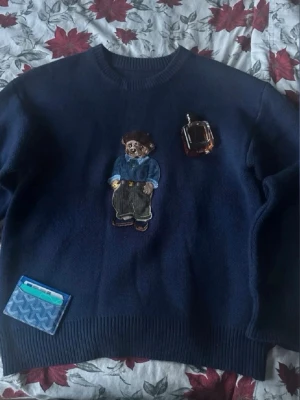 Mörkblå tröja med nallepatch - Mörkblå tröja med rund hals och en stor broderad nallepatch på bröstet. Nallen har basker, blå skjorta och gröna byxor. Tröjan har ribbade muddar vid ärmslut och nederkant. Perfekt för dig som gillar unika detaljer och lekfull stil.